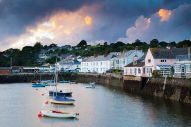 Small boats float by pastel houses along a harbour under a dramatic cloudy sky at sunset.