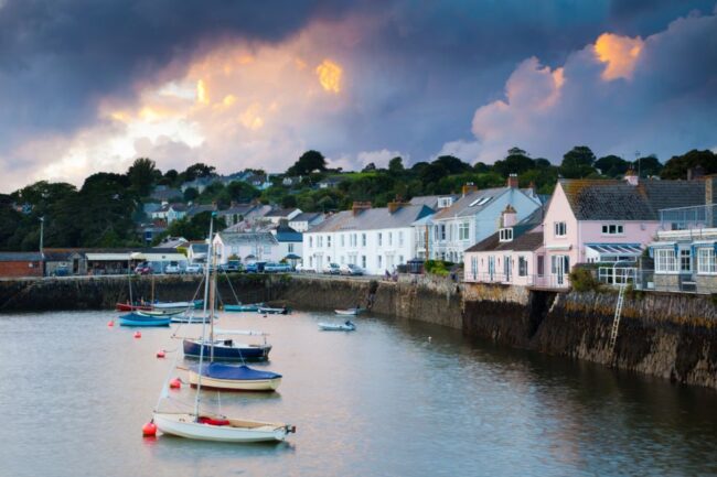 Small boats float by pastel houses along a harbour under a dramatic cloudy sky at sunset.