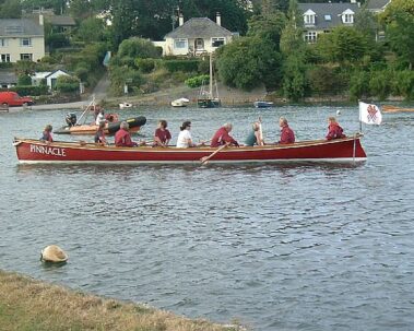 Eight people row a red boat named Pinnacle on a river with houses in the background.
