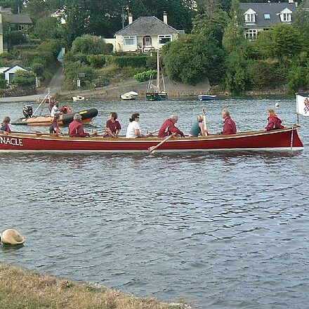 Eight people row a red boat named Pinnacle on a river with houses in the background.