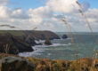 Rocky coastline with cliffs, waves, and grassy foreground under a cloudy blue sky.